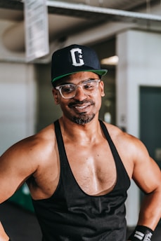 A person wearing a black tank top and a cap with a letter C on it stands in an indoor setting. They have glasses and appear to be smiling slightly.