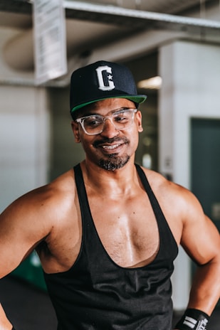A person wearing a black tank top and a cap with a letter C on it stands in an indoor setting. They have glasses and appear to be smiling slightly.