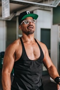 A muscular man wearing a black tank top, glasses, and a black cap is standing in an indoor setting. He appears to be in a gym, given the visible gym equipment in the background. The lighting highlights his physique.