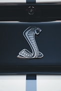 A close-up of a metallic cobra logo on a sleek, glossy surface, possibly the rear of a vehicle. The background is dark, emphasizing the shiny and detailed design of the emblem.