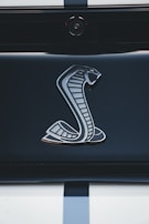 A close-up of a metallic cobra logo on a sleek, glossy surface, possibly the rear of a vehicle. The background is dark, emphasizing the shiny and detailed design of the emblem.