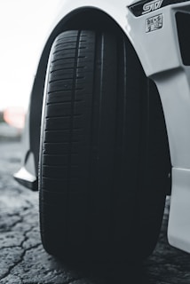 A close-up view of a car tire attached to a white vehicle, with clear tire treads visible. The ground beneath is textured with cracks and the focus is primarily on the tire, with the car's body visible in the background.
