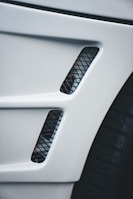 A close-up view of a car's body panel with two vertical rectangular air vents featuring a black mesh pattern, alongside part of a tire visible to the right.