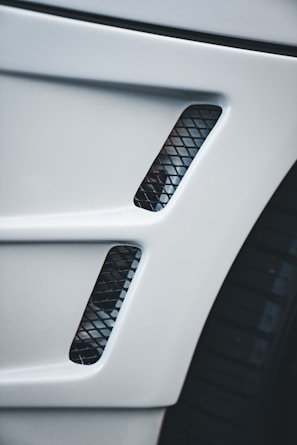 A close-up view of a car's body panel with two vertical rectangular air vents featuring a black mesh pattern, alongside part of a tire visible to the right.