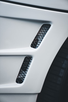 A close-up view of a car's body panel with two vertical rectangular air vents featuring a black mesh pattern, alongside part of a tire visible to the right.