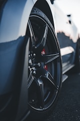 A close-up view of a car wheel featuring sleek, black alloy rims and a visible brake caliper behind the spokes. The tire tread details are prominent, with the car body slightly visible in the background, suggesting a sporty vehicle.