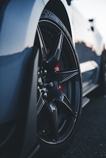 A close-up view of a car wheel featuring sleek, black alloy rims and a visible brake caliper behind the spokes. The tire tread details are prominent, with the car body slightly visible in the background, suggesting a sporty vehicle.