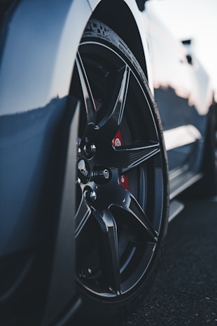 A close-up view of a car wheel featuring sleek, black alloy rims and a visible brake caliper behind the spokes. The tire tread details are prominent, with the car body slightly visible in the background, suggesting a sporty vehicle.