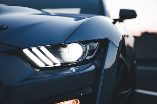 Close-up view of the front headlight of a modern sports car, showcasing sleek design with sharp angles and illuminated lights against a dusk sky background.