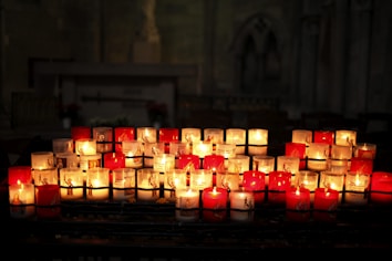 A collection of lit votive candles arranged in multiple rows on a stand inside a dimly lit religious setting. The candles, mostly white and red, have religious imagery and inscriptions on them. The background is blurred, suggesting the setting might be a church or a similar sacred place.