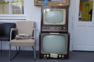 Two vintage televisions are stacked on top of each other next to a beige and a black chair. A stack of newspapers rests on one of the chairs. The setting appears to be indoors with a window and a door visible in the background.
