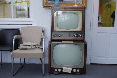 Two vintage televisions are stacked on top of each other next to a beige and a black chair. A stack of newspapers rests on one of the chairs. The setting appears to be indoors with a window and a door visible in the background.