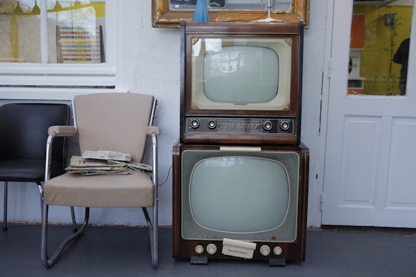Two vintage televisions are stacked on top of each other next to a beige and a black chair. A stack of newspapers rests on one of the chairs. The setting appears to be indoors with a window and a door visible in the background.
