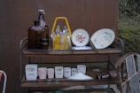 A multi-tier metal cart holds various vintage kitchenware, including brown glass bottles, clear glass bottles with yellow liquid, decorative plates with floral patterns, and cups with floral designs and printed text. The setting suggests an outdoor area with a brown wall and greenery in the background.
