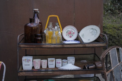 A multi-tier metal cart holds various vintage kitchenware, including brown glass bottles, clear glass bottles with yellow liquid, decorative plates with floral patterns, and cups with floral designs and printed text. The setting suggests an outdoor area with a brown wall and greenery in the background.
