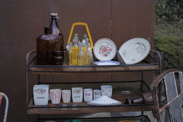 A multi-tier metal cart holds various vintage kitchenware, including brown glass bottles, clear glass bottles with yellow liquid, decorative plates with floral patterns, and cups with floral designs and printed text. The setting suggests an outdoor area with a brown wall and greenery in the background.