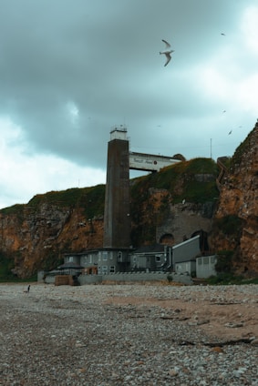 A coastal landscape featuring a tall concrete structure attached to a building set against rugged cliffs. The sky is overcast with several seagulls flying. The beach is rocky with pebbles scattered across the ground.