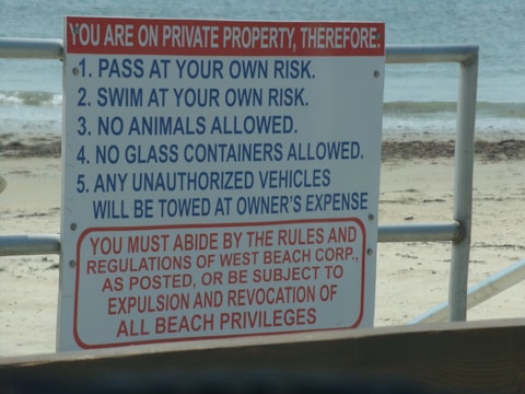 A warning sign lists several rules for a private beach property, detailing regulations such as swimming at your own risk and prohibiting animals and glass containers. The bottom section emphasizes adherence to rules with the potential consequence of being expelled or losing beach privileges.
