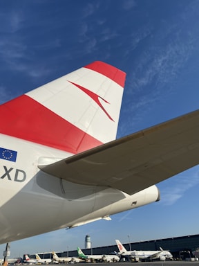 A close-up of an aircraft's tail featuring a red and white design along with the European Union flag emblem. Several other airplanes are parked in the background against a clear blue sky, and a control tower is visible.
