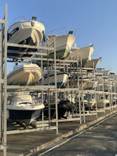 Several motorboats are stored on a large metal rack structure, arranged in rows and stacked vertically. The setting appears to be a marina or boatyard on a sunny day with a clear blue sky.