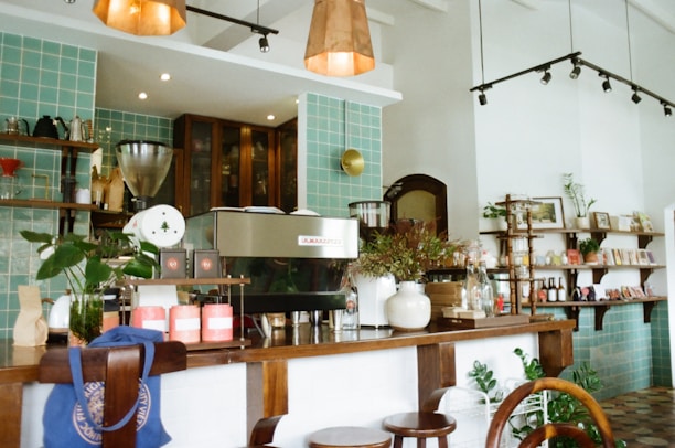 A cozy cafe interior featuring a large coffee machine on a wooden counter surrounded by plants, cups, and assorted decor. Behind the counter, there are shelves with various items including jars, books, and art pieces. The design includes turquoise tiles on the wall, and the ceiling has warm overhead lighting.