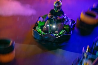 A person wearing a helmet drives a green go-kart on an indoor track. The image is slightly blurred, suggesting a sense of speed. Bright lights cast colorful reflections, creating a dynamic atmosphere.