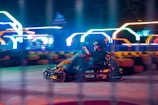 A person driving a go-kart at high speed on a race track with colorful neon lights glowing in the background, creating a dynamic and energetic atmosphere. The motion blur emphasizes the speed and movement of the kart.