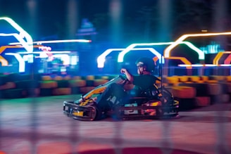 A person driving a go-kart at high speed on a race track with colorful neon lights glowing in the background, creating a dynamic and energetic atmosphere. The motion blur emphasizes the speed and movement of the kart.