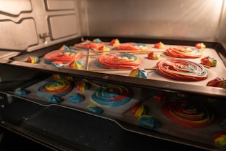 Brightly colored spirals and dollops of whipped confectionery sit on a baking tray inside an oven, with hues of red, blue, yellow, and green visible. Some of the spirals are on sticks, resembling lollipops. The reflections on the oven door glass add depth.