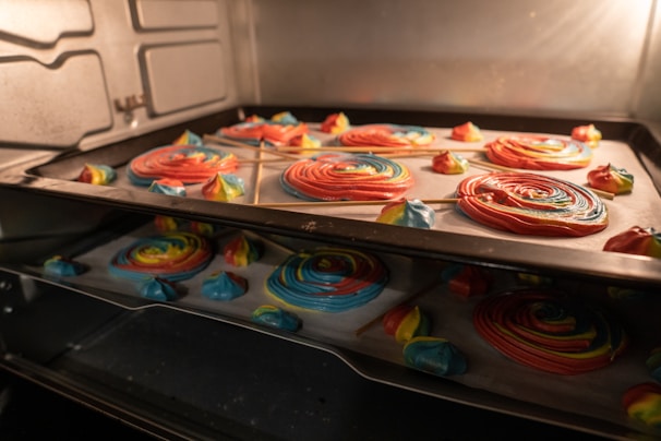 Brightly colored spirals and dollops of whipped confectionery sit on a baking tray inside an oven, with hues of red, blue, yellow, and green visible. Some of the spirals are on sticks, resembling lollipops. The reflections on the oven door glass add depth.