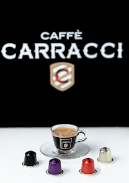 A cup of espresso sits on a clear glass saucer, surrounded by four colorful coffee capsules. The branding reads 'Caffè Carracci' against a stark black background, creating a contrasting visual effect.