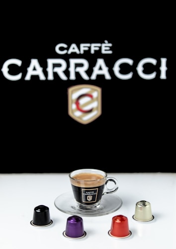 A cup of espresso sits on a clear glass saucer, surrounded by four colorful coffee capsules. The branding reads 'Caff&egrave; Carracci' against a stark black background, creating a contrasting visual effect.