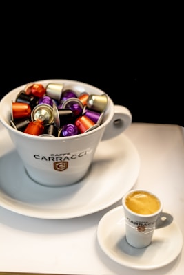 A large white cup filled with various colorful coffee capsules, including shades of purple, orange, and gold, rests on a matching saucer. In the foreground, a small espresso cup with fresh coffee sits on its own saucer. The background is black, creating a stark contrast with the bright colors of the capsules and the white cups.