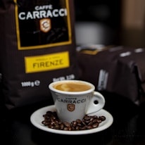 A ceramic cup filled with espresso is placed on a saucer surrounded by coffee beans. In the background, there is packaging with the name 'Caffè Carracci' and the label 'Firenze' visible.