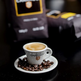 A white espresso cup sits on a matching saucer, filled with crema-topped coffee surrounded by scattered roasted coffee beans. In the background, there are blurred dark coffee bags with labeled branding.