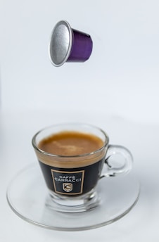 A purple coffee pod appears to be floating above a glass cup filled with espresso, placed on a transparent saucer. The branding on the cup reads 'Caffè Carracci' and the background is plain white.
