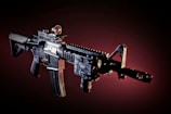An assault rifle is prominently displayed against a dark, gradient background. The rifle features multiple attachments including a scope, a front sight, and a grip. The lighting accentuates the details and contours of the weapon, highlighting its various components.