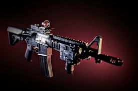 An assault rifle is prominently displayed against a dark, gradient background. The rifle features multiple attachments including a scope, a front sight, and a grip. The lighting accentuates the details and contours of the weapon, highlighting its various components.
