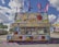 A vibrant carnival stand decorated with colorful images of cotton candy, popcorn, and lemonade. The stand advertises various treats such as candy apples, caramel apples, and popcorn. Several pink and purple flags wave above the stand, and an American flag is displayed on the front. The sky is partly cloudy, and there are additional rides and stalls visible in the background.