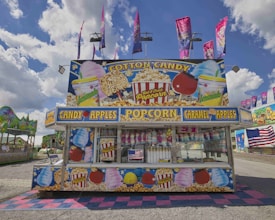 A vibrant carnival stand decorated with colorful images of cotton candy, popcorn, and lemonade. The stand advertises various treats such as candy apples, caramel apples, and popcorn. Several pink and purple flags wave above the stand, and an American flag is displayed on the front. The sky is partly cloudy, and there are additional rides and stalls visible in the background.