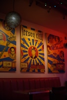 Vibrant poster artwork adorns the wall, showcasing retro-styled illustrations with bold colors and patterns. The images include faces of individuals, guitars, vehicles, and the words 'Jersey City' prominently displayed. The setting is dimly lit, with a cozy atmosphere highlighted by a hanging lamp and a red cushioned bench.