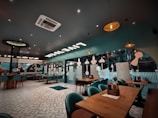 A modern, stylish restaurant interior featuring wooden tables and teal chairs. The ceiling has an array of hanging lights and decorations, with large windows offering a view outside. The ambiance is cozy yet contemporary, with patterned flooring adding a touch of elegance.