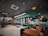 A modern, stylish restaurant interior featuring wooden tables and teal chairs. The ceiling has an array of hanging lights and decorations, with large windows offering a view outside. The ambiance is cozy yet contemporary, with patterned flooring adding a touch of elegance.