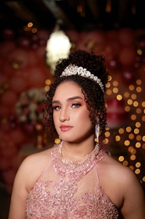 A woman wearing a sparkling tiara and a detailed lace dress poses in a softly lit setting. Her hair is styled in curls, and she is adorned with long, elegant earrings. The background is decorated with pink and golden bokeh lights and balloons, creating a celebratory atmosphere.