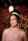A woman wearing a sparkling tiara and a detailed lace dress poses in a softly lit setting. Her hair is styled in curls, and she is adorned with long, elegant earrings. The background is decorated with pink and golden bokeh lights and balloons, creating a celebratory atmosphere.