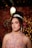 A woman wearing a sparkling tiara and a detailed lace dress poses in a softly lit setting. Her hair is styled in curls, and she is adorned with long, elegant earrings. The background is decorated with pink and golden bokeh lights and balloons, creating a celebratory atmosphere.