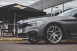 A sleek, dark-colored car is parked in front of a modern building. The vehicle features angular headlights and stylish alloy wheels. There is an outdoor patio umbrella with advertising visible in the background.