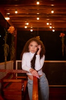 A woman with curly hair is leaning on a wooden railing in a warmly lit outdoor setting. She is wearing a white long-sleeve top and denim jeans, and there are hanging plants and string lights in the background, creating a cozy ambiance.