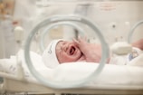 A newborn baby is crying inside an incubator wearing a white hat. The environment appears to be a hospital or neonatal unit, with medical equipment visible and a soft blanket on the incubator bed.
