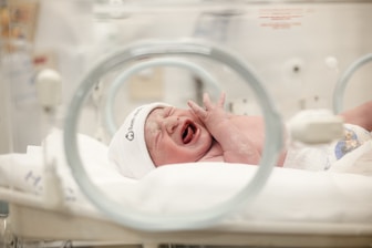 A newborn baby is crying inside an incubator wearing a white hat. The environment appears to be a hospital or neonatal unit, with medical equipment visible and a soft blanket on the incubator bed.
